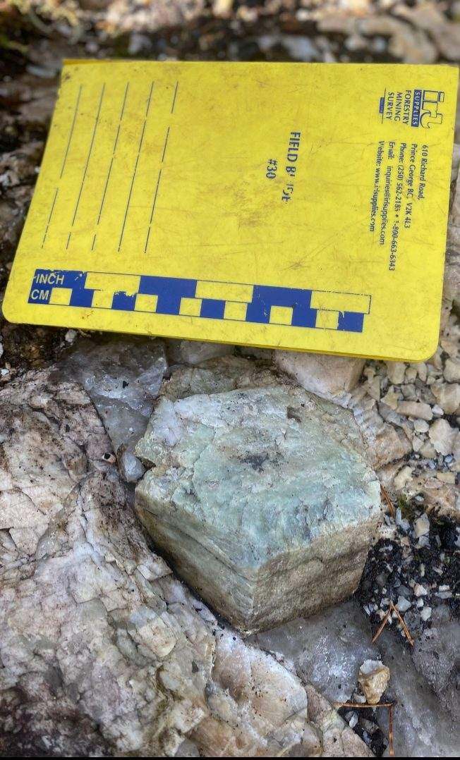 A blocky pale green crystal resting on quartz-rich outcrop beside a yellow field notebook marked with a centimetre and inch scale